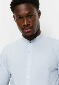 Hellblaues Hemd mit Knopfleiste, das dünne senkrechte Streifen, einen Mandarinkragen und kontrastierende Knöpfe aufweist. Der Stoff wirkt glatt und texturiert.