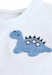 Bílé bavlněné tričko s texturovanou modrou výšivkou dinosaura, černými hroty a bílými kruhovými akcenty. Zapínání na druky u límečku.