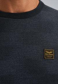 Tmavý textúrovaný sveter s okrúhlym výstrihom, s štvorcovou čiernou a žltou značkovou etiketou na hrudi.