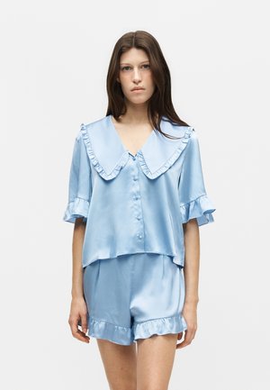 Jeune femme portant une blouse en satin bleu clair à boutons avec un col à volants et un short assorti à volants, debout devant un fond uni.