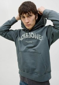 Kapuć sweatshirt u tamnozelenoj boji s teksturiranim prednjim logotipom. Ima kenguru džep i rebraste manžete, izrađen je od mekanog, rastezljivog materijala.