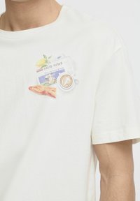 Hvid bomulds t-shirt med et farverigt grafisk design af en avis, kaffekop, citron og croissant på brystet. Korte ærmer, afslappet pasform.