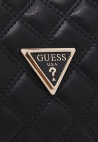 Borsa nera trapuntata con un logo triangolare in oro e nero "GUESS", caratterizzato da un design a punto interrogativo. La texture è liscia, con cuciture visibili.