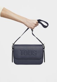 Granatowa skórzana torba na ramię z wytłoczonym logo "TOUS", smukły design, regulowany pasek i srebrne akcenty. Gładka tekstura z delikatnym wzorem.