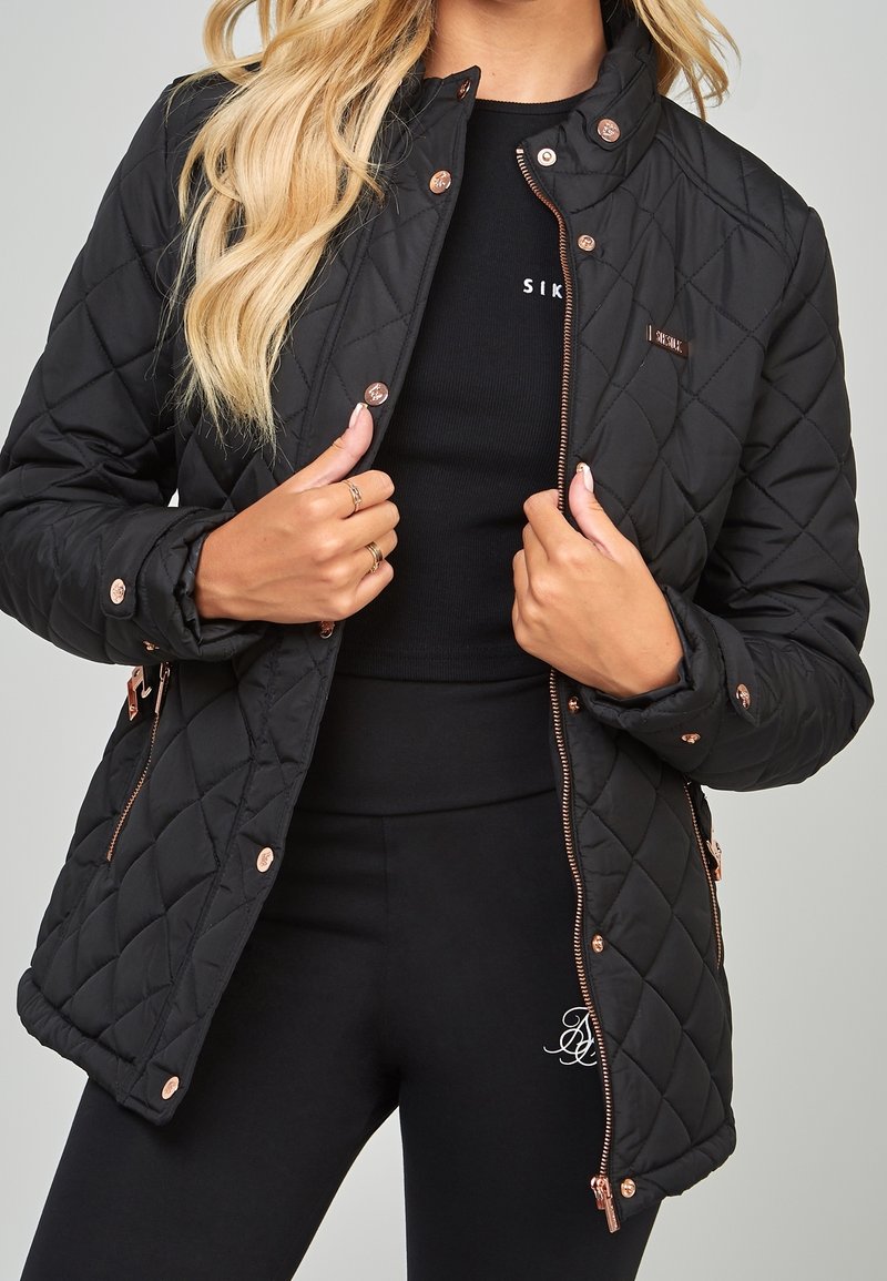 Chaqueta acolchada negra con cuello alto, herrajes en oro rosado y bolsillos laterales, llevada sobre una blusa negra y leggings. Textura suave.