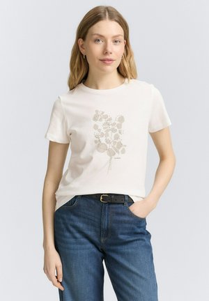 T-shirt crème à manches courtes avec un motif floral dans des tons sourds. Associé à un jean en denim bleu foncé et une ceinture noire.