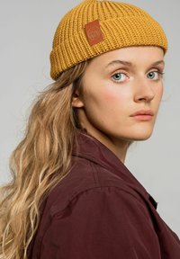 Dzianinowa musztardowa czapka z fakturowanej tkaniny oraz małą brązową skórzaną łatką z logo, noszona przez osobę o falujących włosach i w bordowej kurtce.