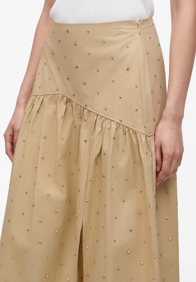 Μπεζ φούστα με διάσπαρτα μικρά μεταλλικά καρφιά, λεπτομέρεια με ραμμένη ραφή, μπροστινή σχισμή, φορεμένη με λευκό ριμπ τοπ μέσα.