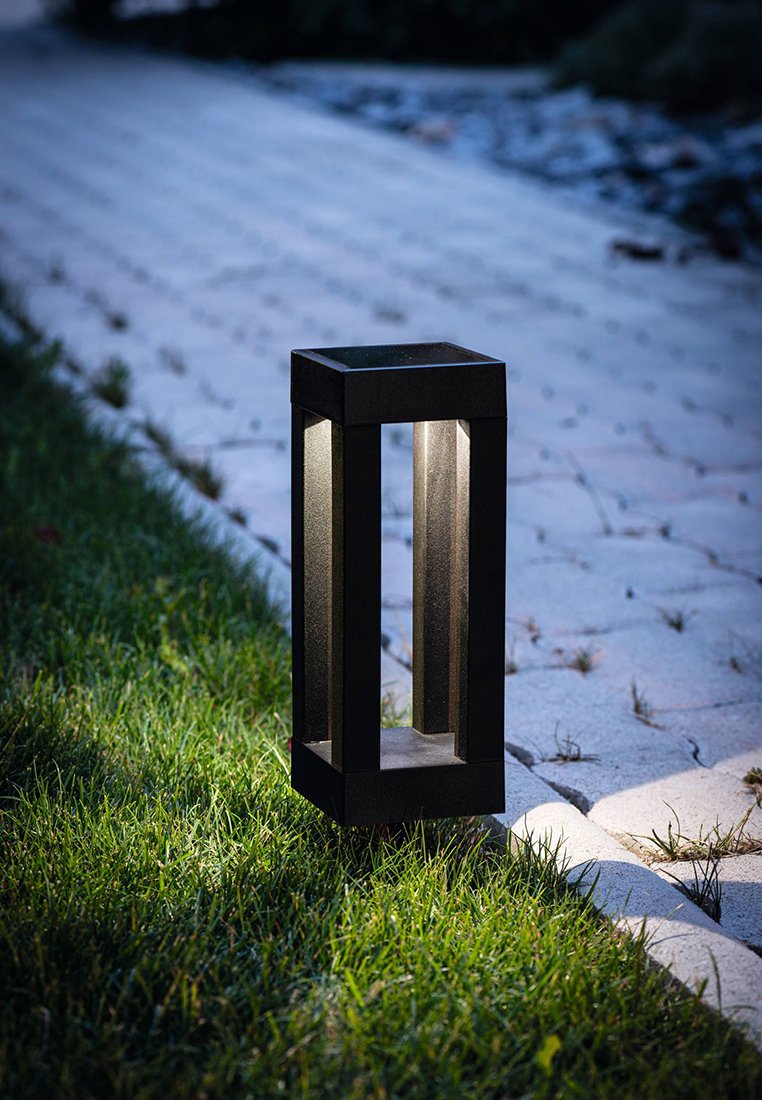 Czarna metalowa lampa zewnętrzna o prostokątnym kształcie, z podświetlonymi bokami, usytuowana na trawie obok kamiennej ścieżki. Z wnętrza emanuje miękkie światło.