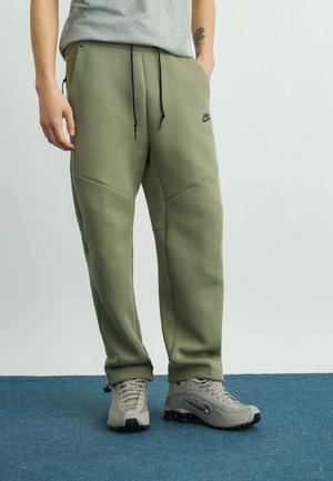 Asmuo dėvintis alyvuogių žalios spalvos Nike sportines kelnes ir šviesiai pilkus Nike sportinius batelius, stovintis mėlyname kilime su viena ranka kišenėje.