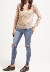 Blusa de tela rosa claro con estampado floral; mangas largas; cuello fruncido. Jeans pitillo azules desgastados; zapatos negros con cordones.