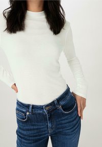 Långärmad, ljus creamfärgad polotröja i kombination med blå denimjeans som har en klassisk femficksdesign och metallknappsstängning.