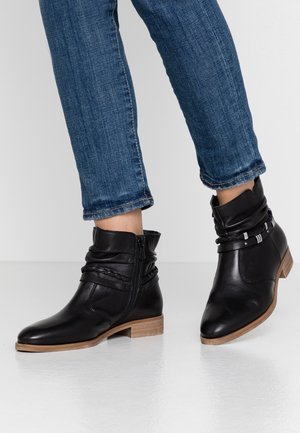 Botas de tobillo de cuero negro con un diseño arrugado, cierre lateral, acento trenzado y tacón de madera, combinadas con jeans azules.
