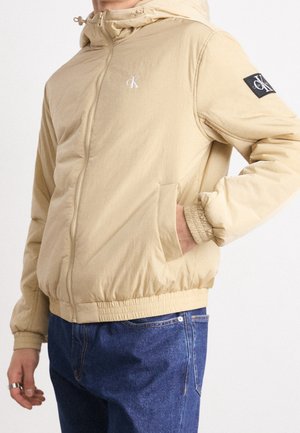 Veste beige zippée avec capuche, poignets et ourlet élastiques, logo Calvin Klein sur la poitrine et la manche, portée avec un jean bleu sur une personne.