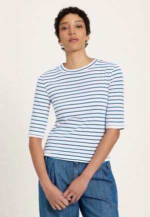 Woman with short curly hair wearing a white and blue striped half-sleeve top and blue high-waisted pants, standing with hands clasped.