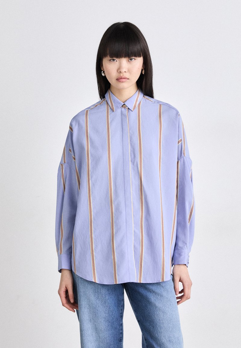 Light blue button-up shirt with vertical orange and cream stripes. Loose fit, wide sleeves, and a classic collar. Paired with light denim jeans.