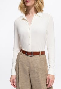 Donna che indossa una camicia bianca a maniche lunghe con bottoni infilata in pantaloni beige a vita alta con una cintura in pelle marrone.