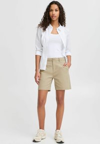 Pantaloni corti in cotone beige con taglio dritto, tasche frontali e passanti per cintura, abbinati a una maglietta senza maniche bianca e una camicia a maniche lunghe. Sneakers bianche.