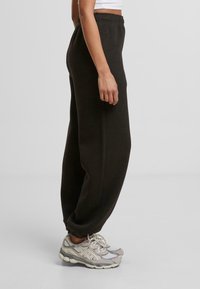 Personne portant un pantalon de survêtement noir avec des poignets élastiques et des chaussures de course gris clair, se tenant de côté contre un fond uni.