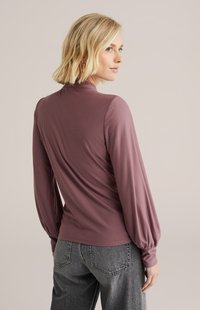 Bluzica dugih rukava u mauve boji s uklopljenim korzetom i naduvanim rukavima, izrađena od mekog, laganog materijala. Ima visok ovratnik i glatku teksturu.