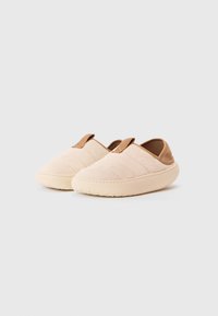 Beige színű, slip-on, párnázott papucsok, suéd textúrával és szövet húzófülekkel fehér háttér előtt.