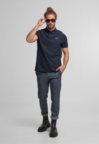 Marineblauwe polo met korte mouwen. Grijze gemusterde broek. Zwarte enkellaarsjes. Houdt een zonnebril vast, informele houding. Eenvoudige accenten en pasvorm.