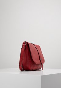 Červená kožená kabelka přes rameno s klopou, nastavitelným popruhem, hladkou texturou a jemným prošíváním podél okrajů, zobrazena z úhlu.
