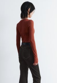 Langärmliges Oberteil in Rostfarbe mit taillierter Passform, kombiniert mit hochgeschnittenen schwarzen Hosen mit Kontrastnähten und Taschen.