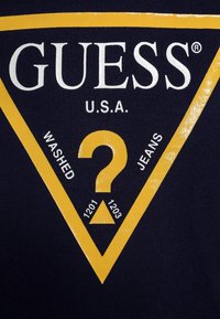 T-shirt in cotone blu navy con un grande grafico triangolare giallo che riporta il testo "GUESS U.S.A." e "WASHED JEANS", insieme a un design con punto interrogativo.
