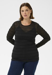 Un haut noir à manches longues avec un tissu texturé superposé et des rayures horizontales, accompagné d'un jean bleu foncé.