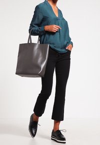 Donna in blusa teal e pantaloni neri a vita alta, che tiene una grande borsa grigia e indossa scarpe con plateau nere, in piedi davanti a uno sfondo bianco.