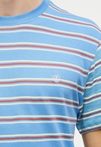 Μπλε μπλουζάκι με οριζόντιες ανοιχτές κίτρινες, λευκές και σκουριασμένες ρίγες και μικρό λευκό κεντημένο λογότυπο στο στήθος, που φοριέται από άτομο.