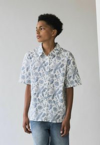 Camisa de botón de manga corta en blanco con estampado floral azul, que presenta un cuello extendido y un ajuste relajado, combinado con unos jeans de denim claros.