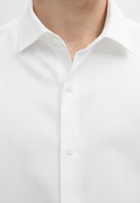 Close-up van een persoon die een wit overhemd draagt met een puntige kraag en twee zichtbare knopen dicht bij de hals vastgeknopen.