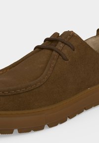 Chaussure décontractée en daim marron avec une semelle épaisse en caoutchouc et des lacets marron foncé, avec des coutures visibles le long du bord supérieur.