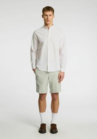 Camisa blanca de botones con cuello mao, combinada con pantalones cortos verde claro. Zapatos marrones y calcetines blancos completan el look.
