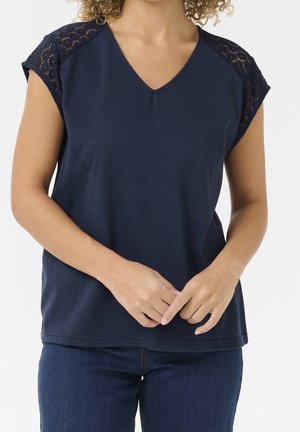 Nainen, joka tekee tummansinisen V-kaula-aukoisen topin pitsisillä lyhyillä hihoilla ja tummansinisillä farkuilla, kädet hellästi ristissä edessään.