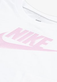 Vit bomullst-shirt med en stor rosa glittrande Nike-logo, rund halsringning och mjuk textur. Närbild visar logotypens detaljer.