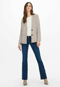 Světle béžový blazer s černými knoflíky, nošený přes bílý top. Kombinovaný s modrými rozšířenými džíny. Hladká látka a dobře padnoucí střih.