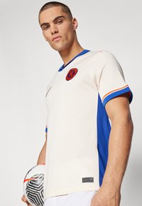 Ljus beige tröja med blå sidopaneler och orange detaljer, med rund hals, korta ärmar och en laglogo på bröstet.