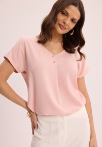 Różowa bluzka, gładki materiał, krótkie zwinięte rękawy, dekolt w serek z trzema guzikami, luźny krój, zestawiona z białymi spodniami i złotą bransoletką.