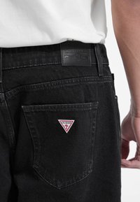 Blugi negri din denim cu un eticheta din piele Guess la talie și o patch triunghiulară roșie pe buzunarul din spate.