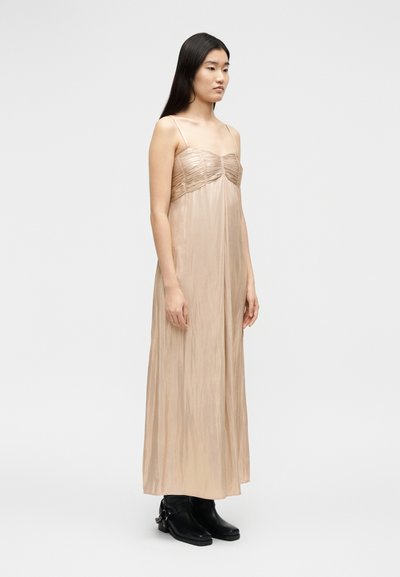 Γυναίκα με μακριά μαύρα μαλλιά που φοράει μακρύ μπεζ φόρεμα χωρίς τιράντες και μαύρα μποτάκια μέχρι τον αστράγαλο, στέκεται μπροστά σε απλό φωτεινό φόντο.