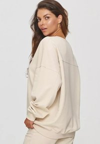 Beżowy, oversize sweter z okrągłym dekoltem, opadającymi ramionami, prążkowanymi mankietami i dołem. Gładki, miękki materiał z minimalnymi detalami.