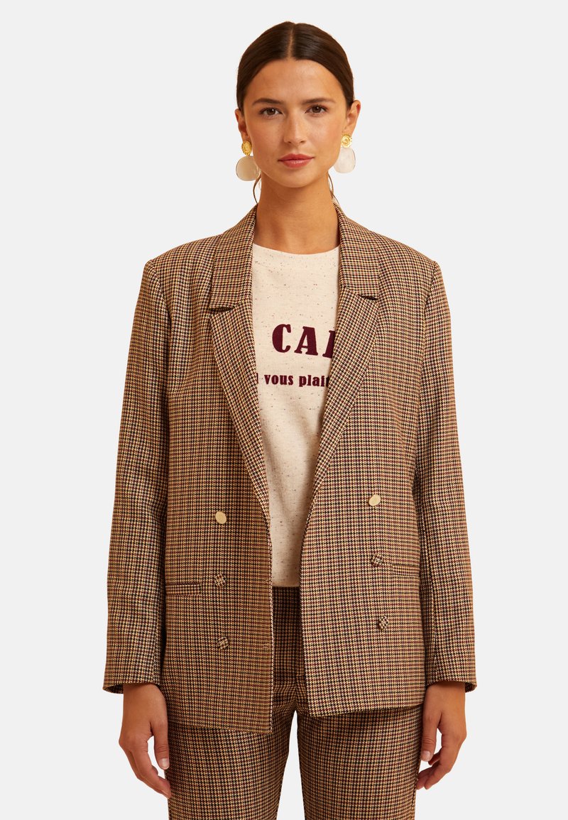 Blazer de doble botonadura a cuadros marrones con botones dorados, que presenta bolsillos y un corte relajado, llevado sobre una camiseta gráfica de color claro.