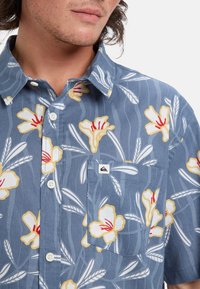 Košile na knoflíky s krátkým rukávem v modré barvě, s květinovým vzorem žlutých a bílých květin. Obsahuje náprsní kapsu s logem.