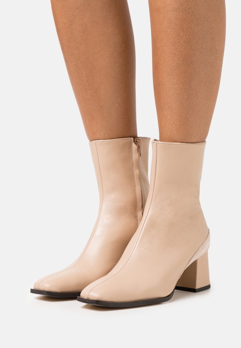 Bottes en cuir beige montant jusqu'au milieu du mollet, avec talons carrés et fermetures éclair latérales, portées sur des jambes nues sur un fond uni.