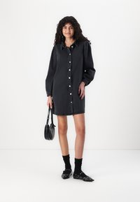 Rochie din denim negru cu butoni, mâneci lungi și o croială ușor largă. Asociată cu șosete negre și pantofi negri, plus o poșetă mică neagră.