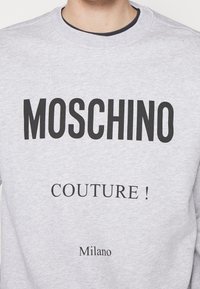 Šedá mikina s velkým černým nápisem „MOSCHINO“ nahoře, pod ním „COUTURE !“ a na dně „Milano“. Design s kulatým výstřihem.
