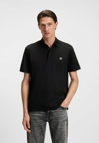 Czarna koszulka polo z kołnierzykiem, krótkimi rękawami i okrągłym logo na klatce piersiowej. Tkanina wydaje się gładka; zestawiona z jasnoszarymi dżinsami.